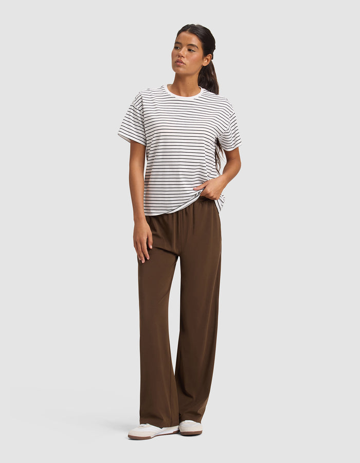 A woman stands against a plain background wearing Cozy Earth Women's Studio Wide Leg Pants in brown, a white and black striped short-sleeve t-shirt, and white slip-on shoes. Her long dark hair is pulled back, and her left hand is in her pocket.|Color:Espresso