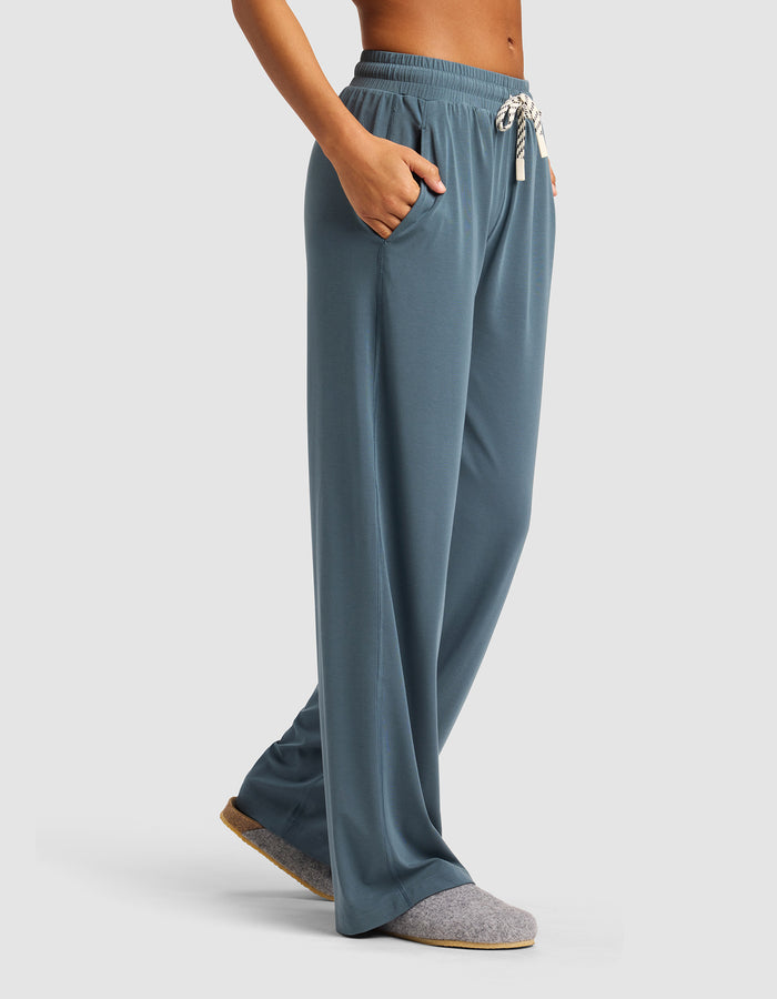 A woman stands with hands in the pockets of Cozy Earth's Women's Studio Wide Leg Pant—loose-fitting, blue drawstring lounge pants—paired with gray slippers against a plain, light background.|Color:Dawn