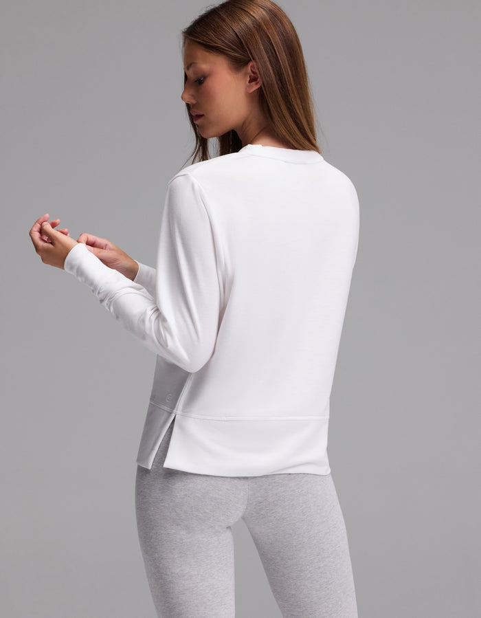 A woman with long brown hair is standing sideways against a plain gray background, wearing the Cozy Earth Women’s StudioLite Crewneck in white and light gray leggings, while looking at her hand.|Color:White