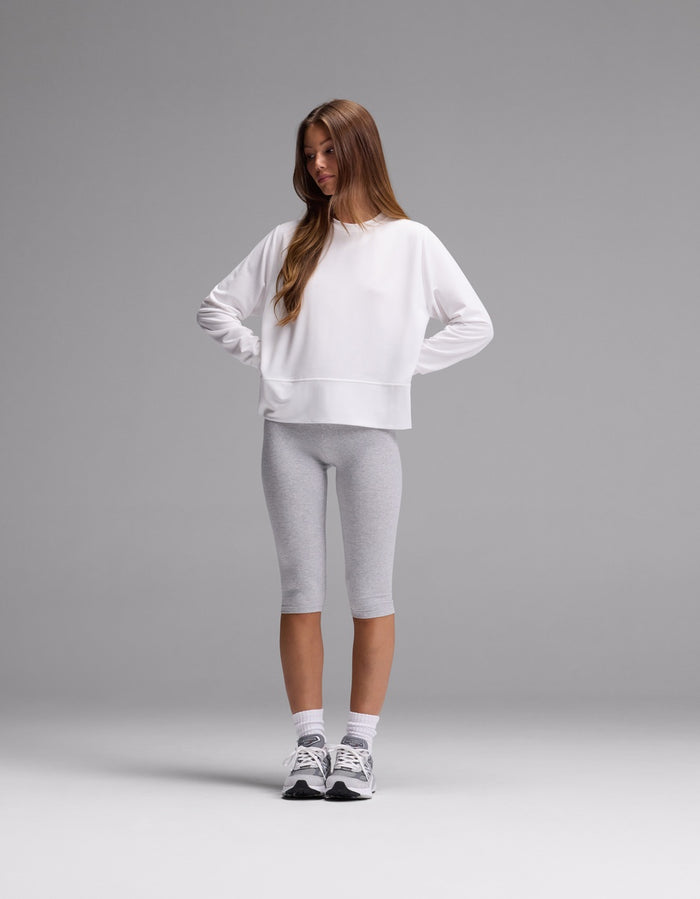 A young woman stands with hands on hips, wearing the Cozy Earth Women’s StudioLite Crewneck in white, paired with light gray knee-length leggings, white socks, and gray sneakers against a plain gray background.|Color:White