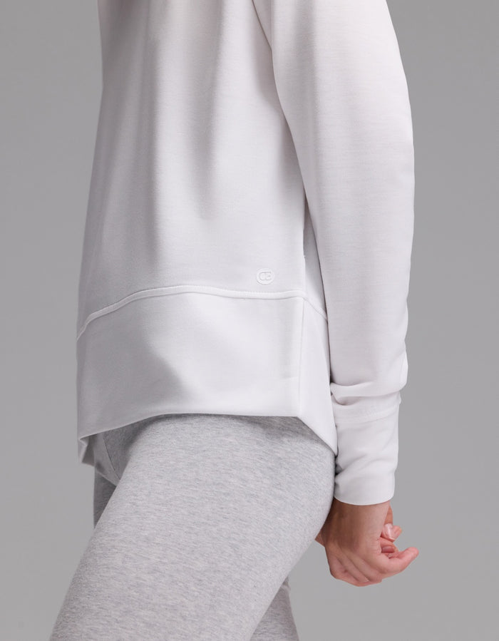 A woman stands sideways against a plain gray background, wearing the Cozy Earth Women’s StudioLite Crewneck in white and light gray leggings, with one arm relaxed at her side.|Color:White