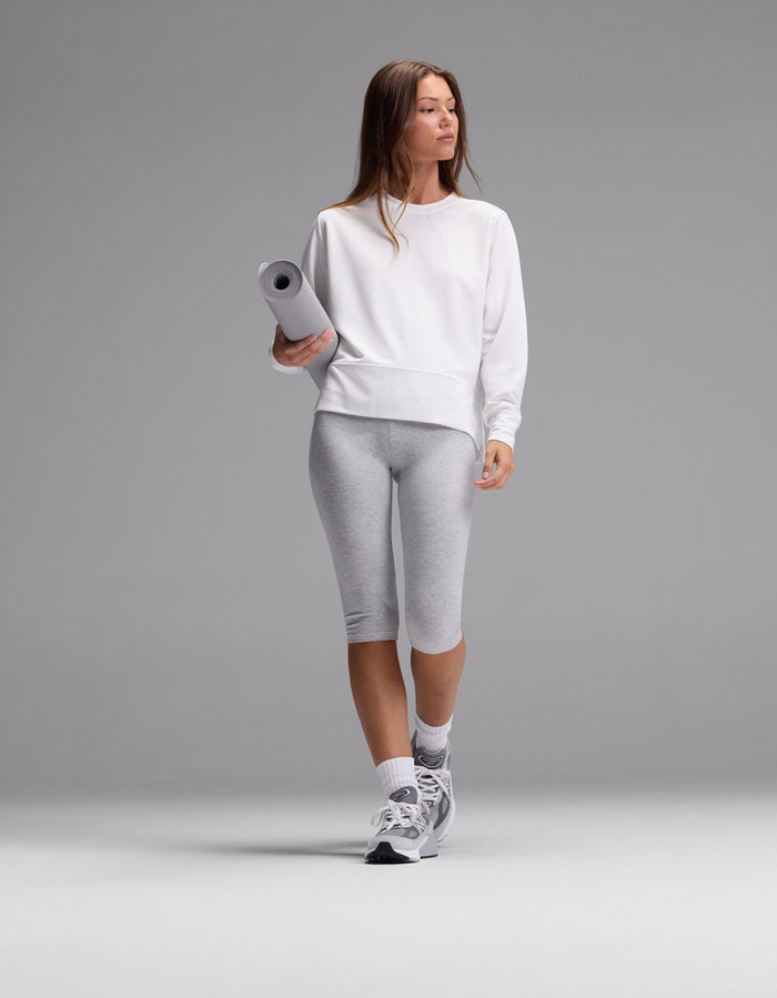 A woman wearing the Cozy Earth Women’s StudioLite Crewneck, paired with gray capri leggings and sneakers, holds a rolled yoga mat while standing against a plain gray background.|Color:White