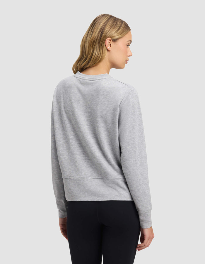 A woman with shoulder-length blonde hair faces away, wearing the Cozy Earth Women's StudioLite Crewneck in light gray with black leggings, set against a plain light gray background.|Color:Heather Grey