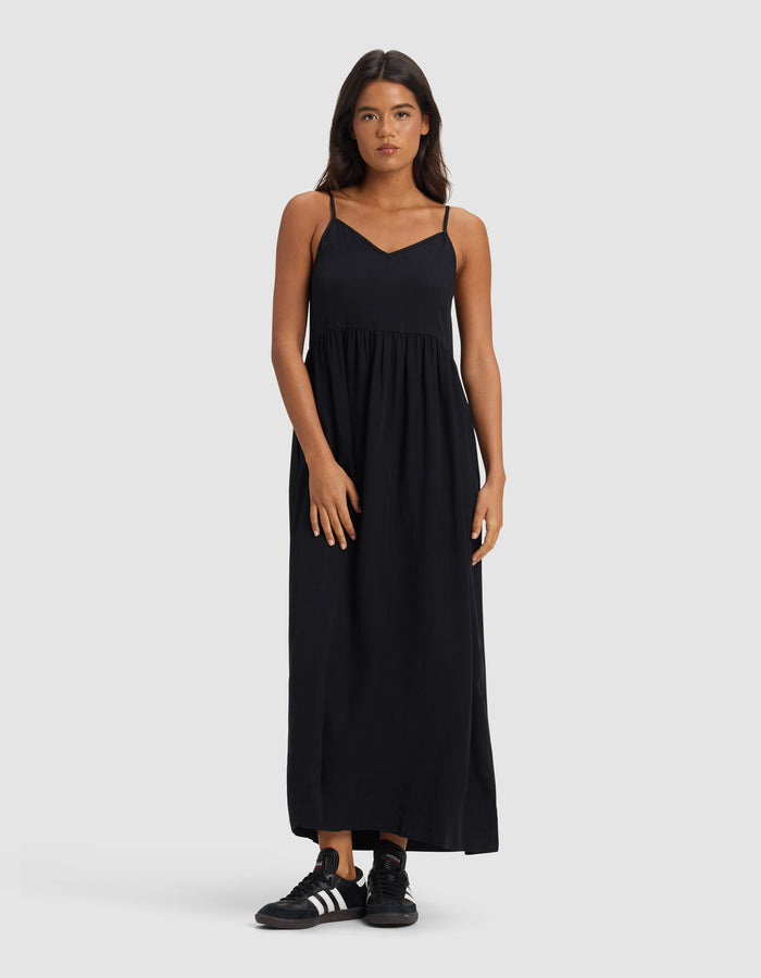 A woman faces forward in a Cozy Earth Women's Sunset Gathered Dress – Last Chance, an ankle-length, sleeveless black dress with thin straps, paired with black Adidas sneakers against a plain light gray background.|Color:Jet Black