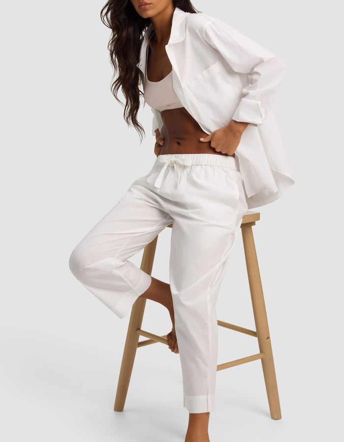 A woman with long wavy hair sits on a wooden stool against a plain light background, wearing Cozy Earth's Women's Sutton Cropped Pajama Pant paired with a white button-up shirt and crop top.|Color:White
