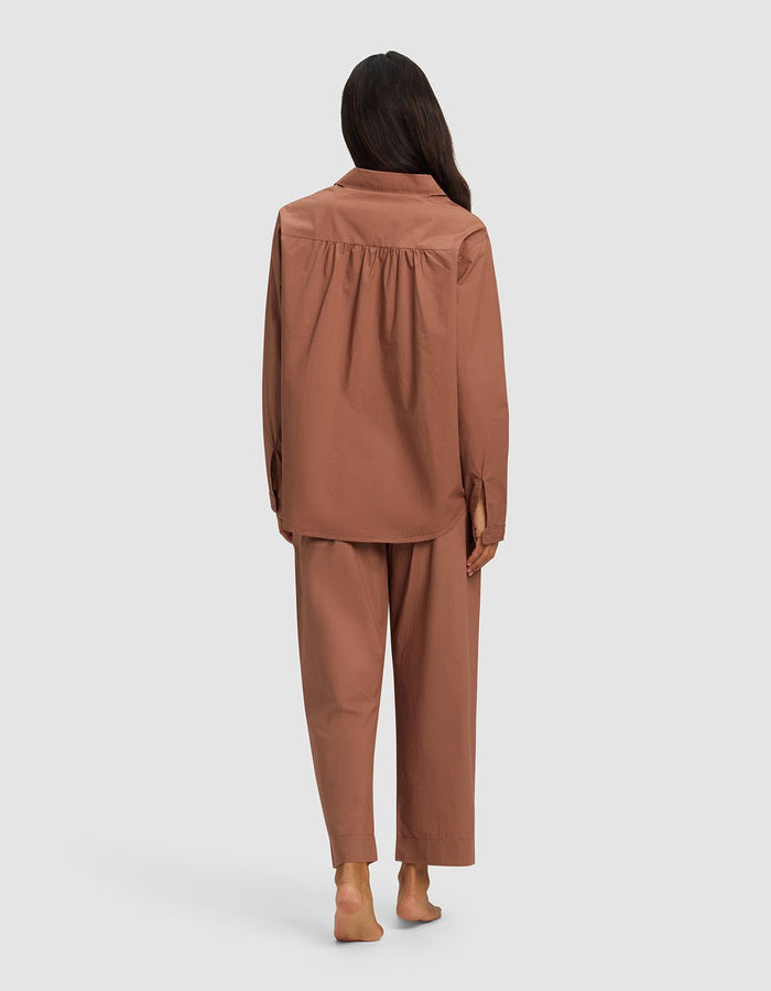 A barefoot woman with long dark hair is shown from behind wearing the Cozy Earth Women's Sutton Long Sleeve Pajama Set in brown, featuring a loose fit and long sleeves and pants, against a plain light gray background.|Color:Saddle
