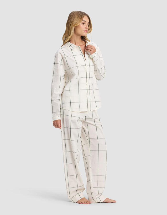 A woman stands barefoot, wearing the Cozy Earth Women's Sutton Long Sleeve Pajama Set in white with thin black grid lines, featuring a button-up shirt and loose pants. She looks to the side and touches her chin.|Color:Bone Windowpane