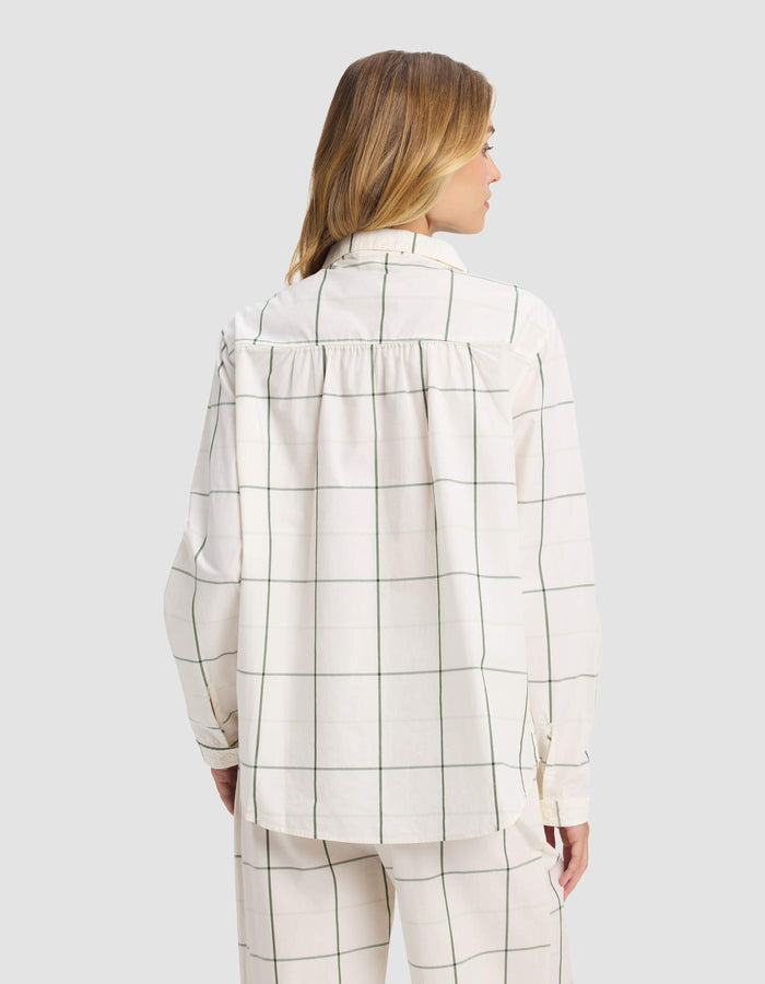 A woman with light brown hair faces away from the camera, wearing the Cozy Earth Women's Sutton Long Sleeve Pajama Top—a loose-fitting, white shirt with a green windowpane grid pattern—along with matching pants.|Color:Bone Windowpane