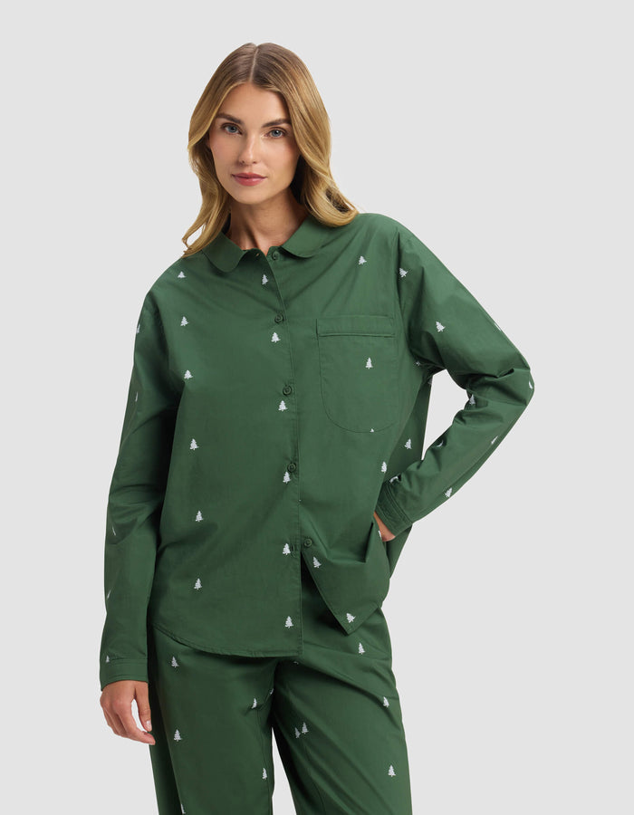 A woman models the Cozy Earth Women's Sutton Long Sleeve Pajama Top, featuring a matching green set with a small white tree pattern, as she poses with one hand in her pocket against a light gray background.|Color:Jade Tree