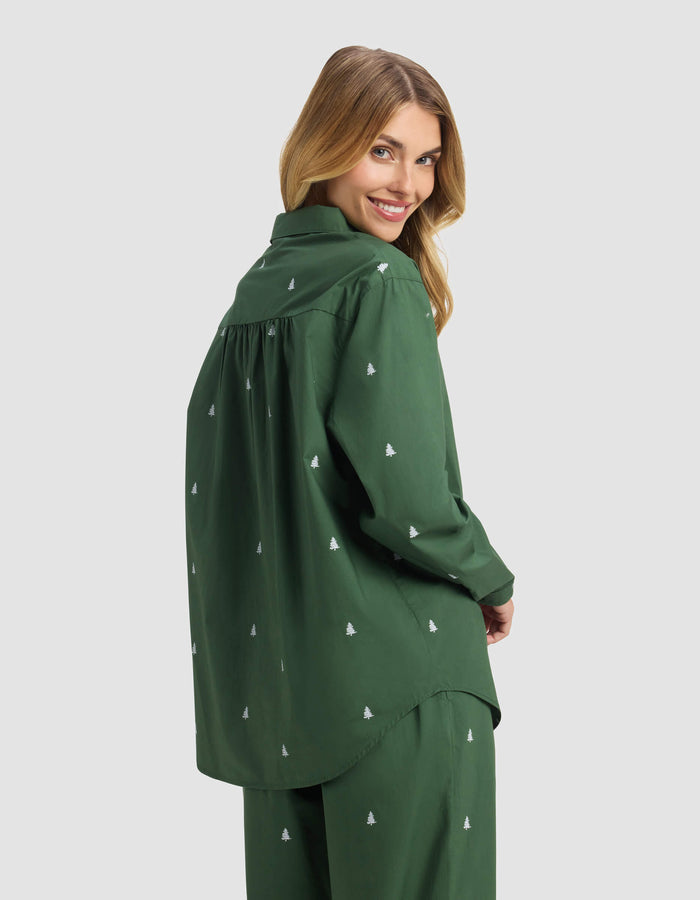 A woman with long blonde hair smiles over her shoulder, wearing the Cozy Earth Women's Sutton Long Sleeve Pajama Top in dark green, paired with matching pants featuring small white embroidered tree patterns. The background is light gray.|Color:Jade Tree