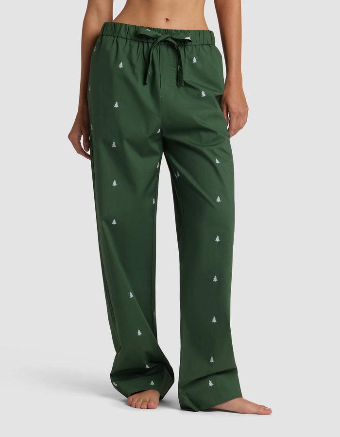 A person stands barefoot against a plain background wearing Cozy Earth’s Women’s Sutton Pajama Pant in dark green, featuring a drawstring waist and small white tree patterns. The loose-fitting pants reveal the person’s midriff and feet.|Color:Jade Tree