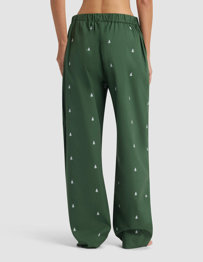 A woman models the Women's Sutton Pajama Pant by Cozy Earth, shown from the back waist down in loose-fitting dark green fabric with a subtle white tree pattern against a plain light background.|Color:Jade Tree
