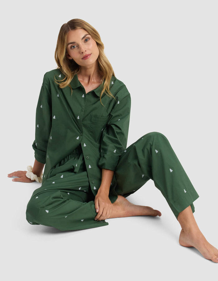 A woman with long blonde hair sits barefoot on the floor in Cozy Earth’s Women’s Sutton Pajama Pant—a dark green set with small white tree patterns—against a plain light gray background.|Color:Jade Tree