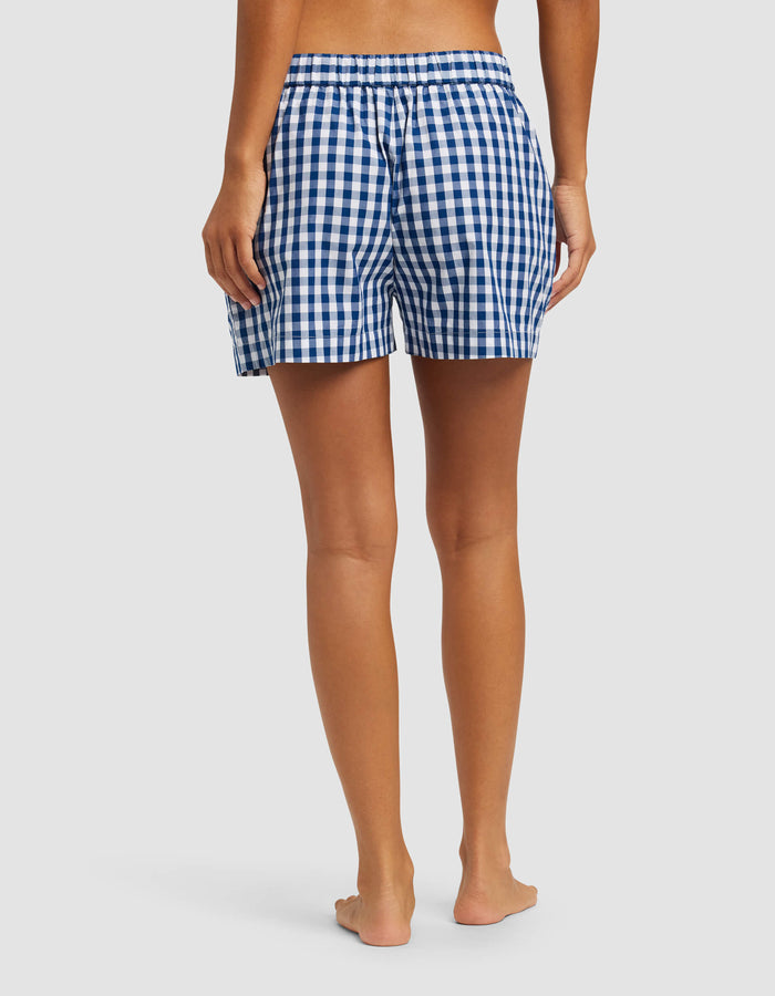 A person stands barefoot facing away, wearing Cozy Earth's Women's Sutton Pajama Short—blue and white checkered with an elastic waistband—against a plain light background.|Color:Pacific Gingham