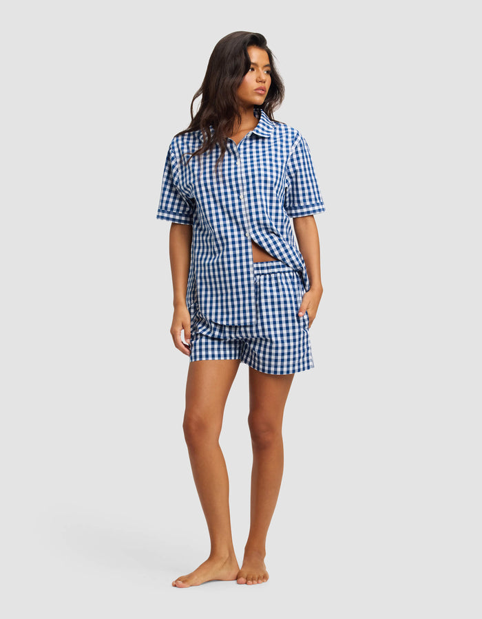 A woman stands barefoot against a plain light background wearing Cozy Earth's Women's Sutton Pajama Shorts set in blue and white gingham, featuring a button-up short-sleeve shirt and matching shorts.|Color:Pacific Gingham