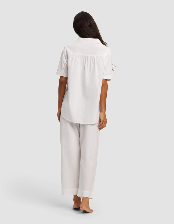 A woman stands barefoot, facing away, wearing the Cozy Earth Women's Sutton Short Sleeve & Pant Pajama Set in white against a plain light background.|Color:White