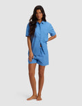 A woman with long dark hair stands barefoot, posing with one hand on her hip against a light gray background, wearing the Women's Sutton Short Sleeve Pajama Set by Cozy Earth—a matching light blue button-up shirt and shorts.|Color:Sailor