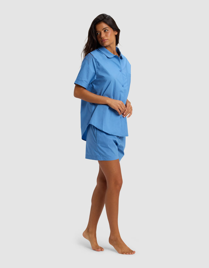 A woman stands barefoot on a light gray background, looking to her left, wearing Cozy Earth's Women's Sutton Short Sleeve Pajama Set—a loose-fitting blue button-up shirt with matching blue shorts.|Color:Sailor