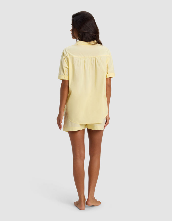 A woman with long dark hair stands barefoot, facing away, wearing the Cozy Earth Women's Sutton Short Sleeve Pajama Set in light yellow against a plain light gray background.|Color:Meringue