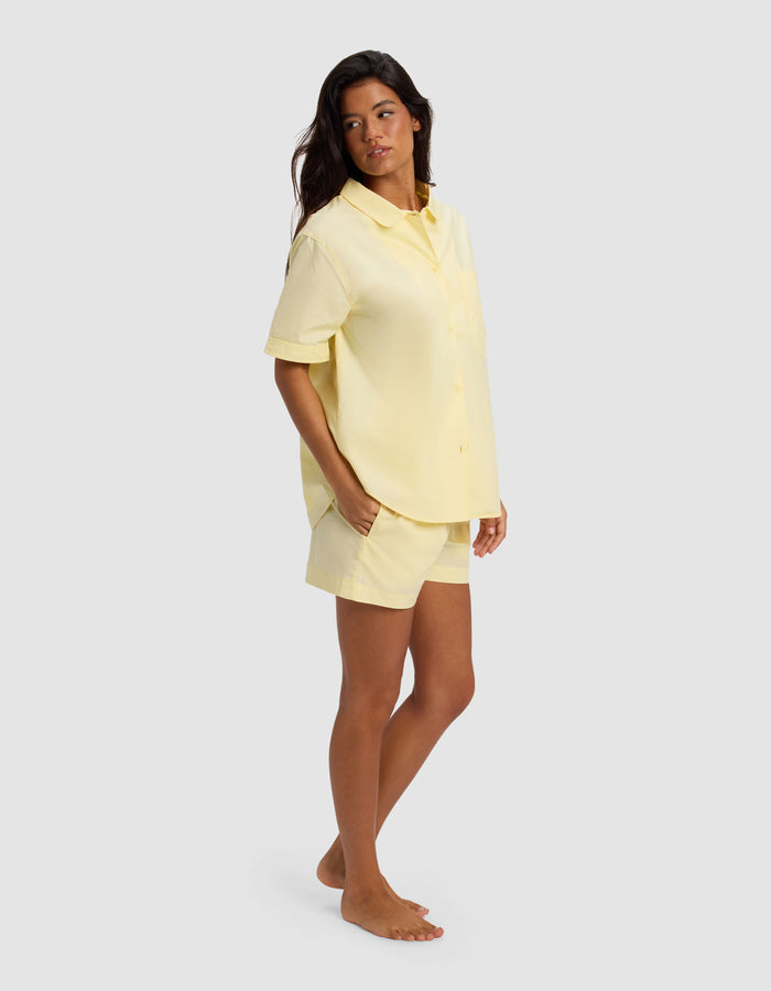 A woman stands barefoot against a light gray background, wearing the Cozy Earth Women's Sutton Short Sleeve Pajama Set in yellow, with her hands in her pockets and looking slightly to the side.|Color:Meringue