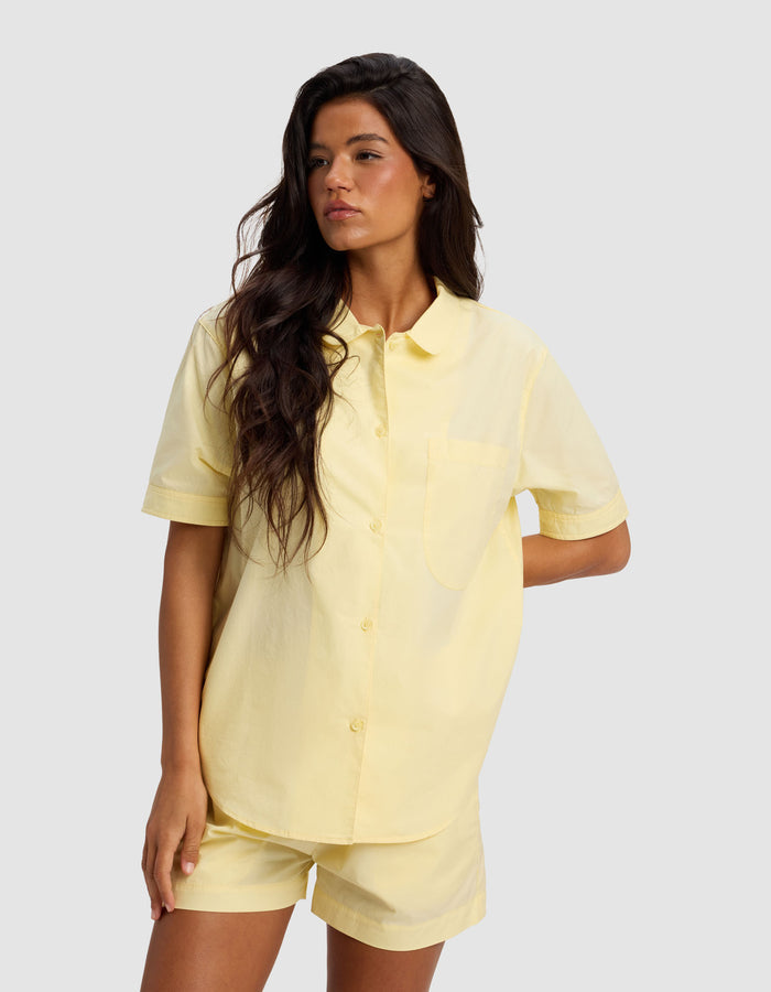 A woman with long dark hair wears the Cozy Earth Women's Sutton Short Sleeve Pajama Top in light yellow, paired with matching shorts, standing against a plain light gray background.|Color:Meringue