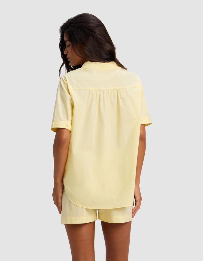 A woman with long dark hair is seen from behind, wearing the Cozy Earth Women's Sutton Short Sleeve Pajama Top in pale yellow, paired with matching shorts, standing against a light gray background.|Color:Meringue