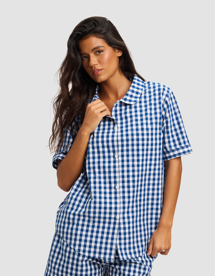 A woman with long dark hair wears the Cozy Earth Women's Sutton Short Sleeve Pajama Top, featuring a blue and white checkered pattern, standing against a plain light gray background.|Color:Pacific Gingham