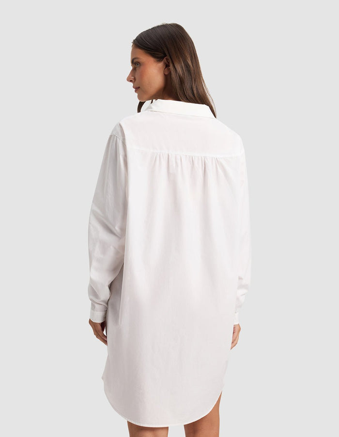A woman with long brown hair faces away, wearing the Cozy Earth Women's Sutton Sleep Dress—a loose-fitting white, long-sleeve shirt dress—set against a plain light gray background.|Color:White