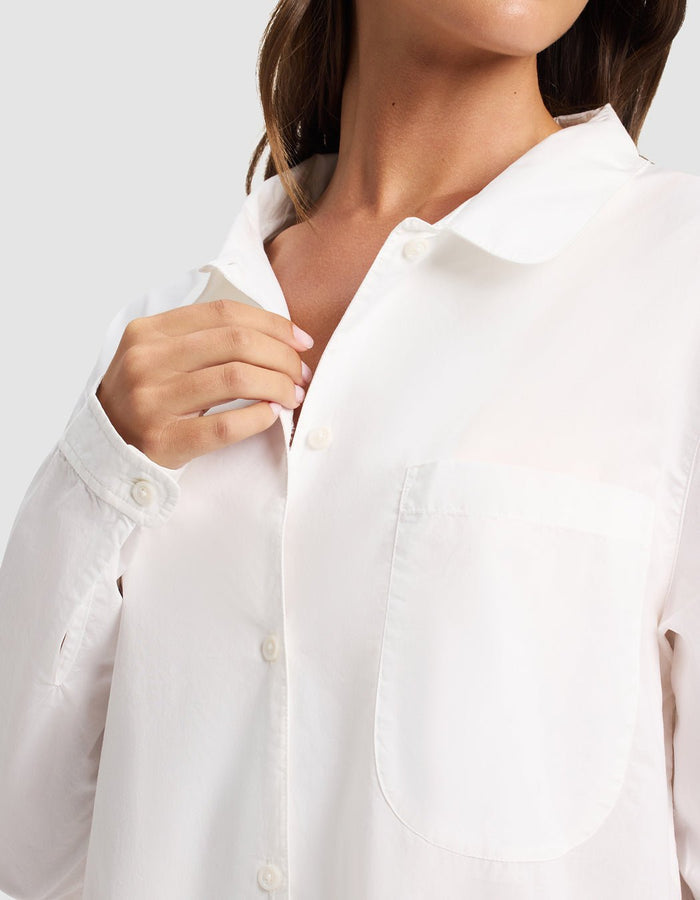 A woman wearing the Cozy Earth Women's Sutton Sleep Dress, a white button-up style with a front pocket, touches the collar with one hand. The image shows her from shoulders to mid-torso against a neutral background.|Color:White