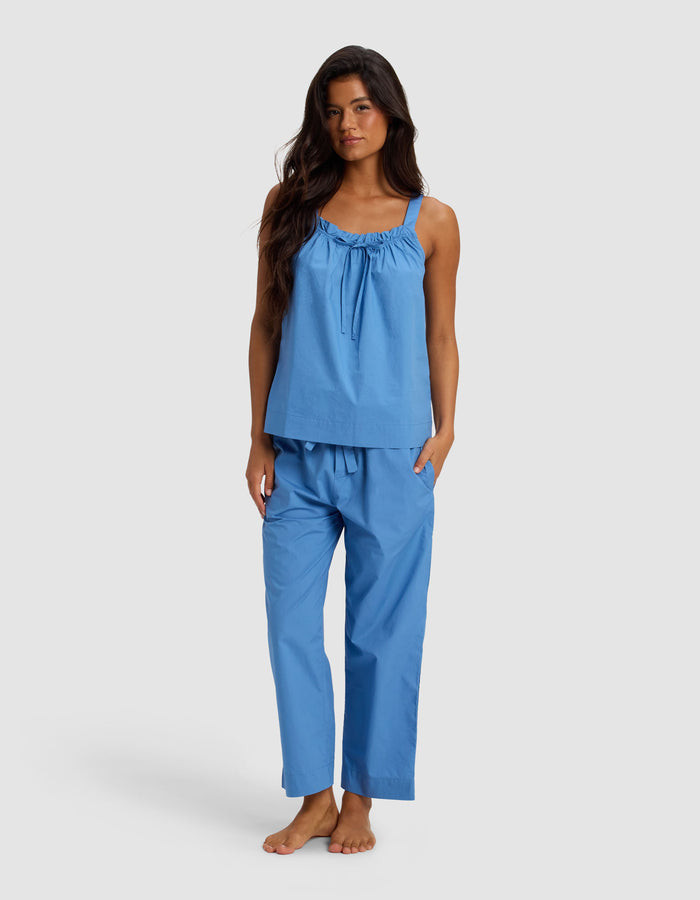 A woman with long dark hair stands barefoot against a plain light background, wearing the Cozy Earth Women's Sutton Tank & Cropped Pant Pajama Set in light blue.|Color:Sailor