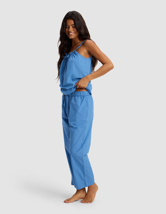 A woman with long dark hair stands barefoot, smiling in the Cozy Earth Women's Sutton Tank & Cropped Pant Pajama Set—a loose-fitting blue sleeveless top with thin straps and matching pants—against a plain light background.|Color:Sailor