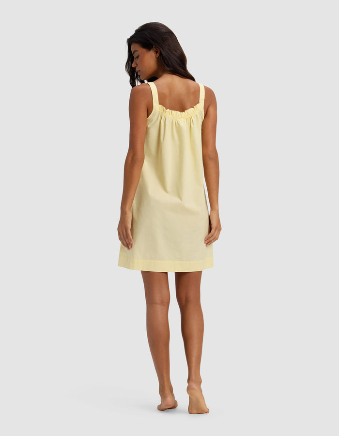 A woman with long dark hair stands barefoot, facing away, wearing the Cozy Earth Women's Sutton Tank Sleep Dress in light yellow, featuring a sleeveless, loose fit and gathered upper back detail, against a plain light gray background.|Color:Meringue
