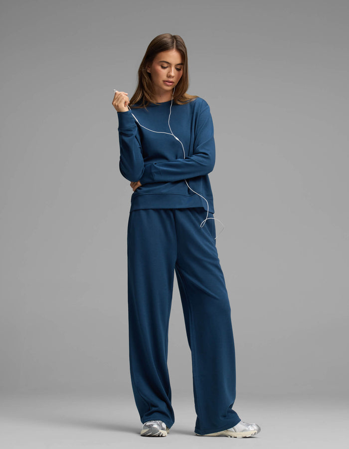 A woman wears the Cozy Earth Women's Versa Crewneck & Straight Leg Pant Set with sneakers, standing against a gray background and holding wired earphones with a relaxed expression.|Color:Obsidian