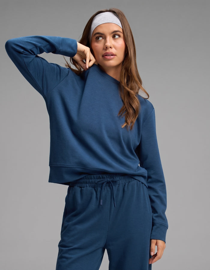A woman stands against a plain background, wearing the Cozy Earth Women's Versa Crewneck & Wide Leg Pant Set in blue, paired with a light gray headband. She raises one arm to touch her face.|Color:Obsidian
