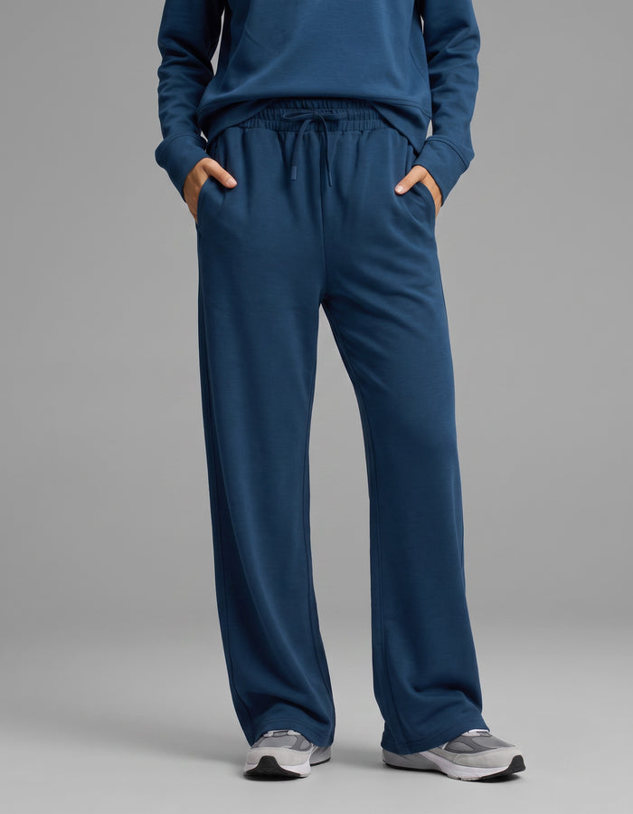 A person is shown from the waist down wearing the Cozy Earth Women's Versa Crewneck & Wide Leg Pant Set in blue, with hands in the pockets of the wide leg pants. They are also wearing gray athletic shoes, standing against a plain light gray background.|Color:Obsidian