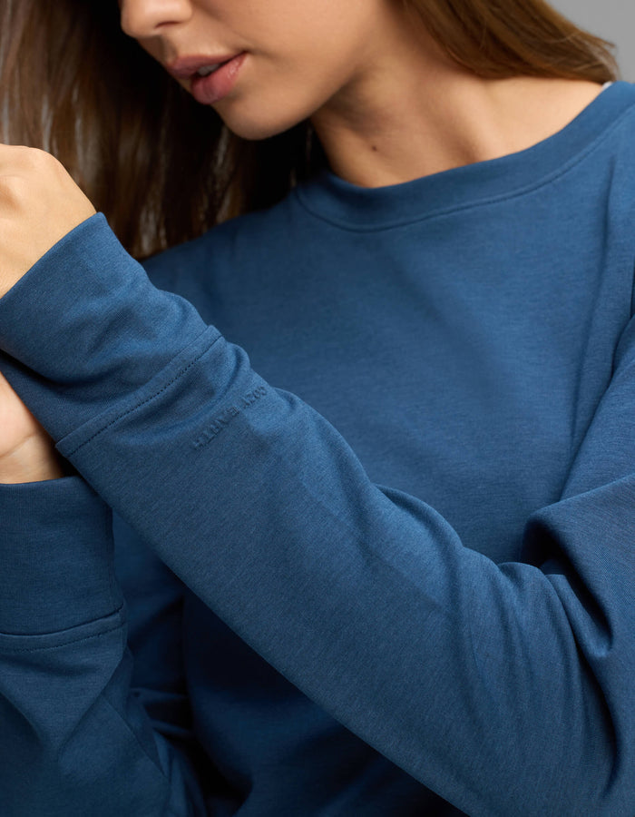 A woman wearing the Cozy Earth Women's Versa Crewneck in blue, featuring subtle embroidered text on the sleeve, holds her hands together near her face. Her head is turned slightly to the side, and only part of her face is visible.|Color:Obsidian