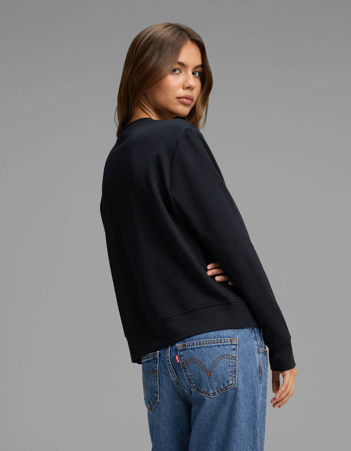 A woman with long brown hair wears the Cozy Earth Women's Versa Crewneck in black and blue jeans, standing with her back turned and head looking over her shoulder against a plain gray background.|Color:Jet Black