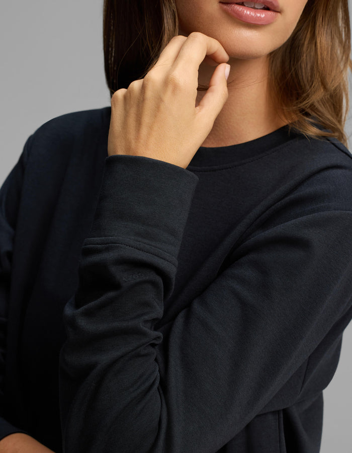 A woman wearing the Cozy Earth Women's Versa Crewneck in black rests her chin on her hand, with only the lower half of her face and upper torso visible.|Color:Jet Black