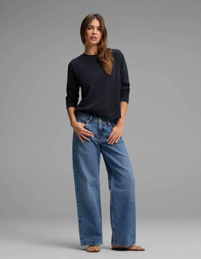 A woman with long brown hair stands against a gray background wearing the Cozy Earth Women's Versa Crewneck, paired with loose blue jeans and sandals. She poses with one hand in her pocket and the other resting by her side.|Color:Jet Black