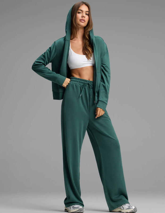 A woman stands confidently against a plain gray background, wearing Cozy Earth's Women's Versa Zip-Down Hoodie & Straight Leg Pant Set in green, paired with a white sports bra and sneakers.|Color:Uniform