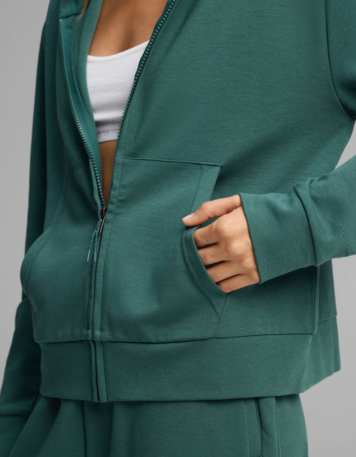 A person wearing the Cozy Earth Women's Versa Full-Zip Hoodie in green with their right hand in the front pocket; a white top is partially visible underneath.|Color:Uniform
