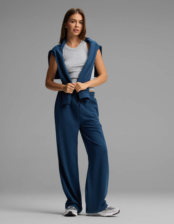 A woman stands against a plain gray background, looking forward with a relaxed posture, wearing Cozy Earth's Women's Versa Straight Leg Pant in blue, paired with a gray tank top and a matching blue sweatshirt draped over her shoulders.|Color:Obsidian