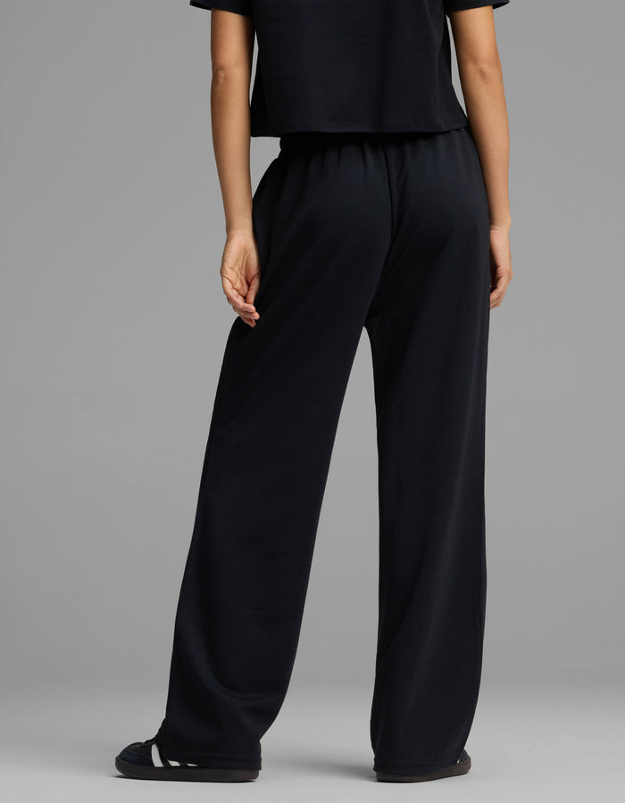 A person stands facing away, wearing Cozy Earth's Women's Versa Straight Leg Pant in black. The background is plain gray, and only the lower half of their body is visible.|Color:Jet Black