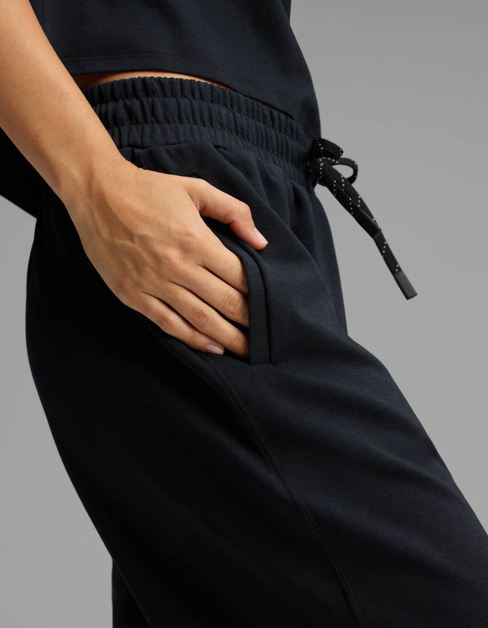 A person wearing Cozy Earth's Women's Versa Straight Leg Pant in black, paired with a matching black top, has their right hand in the pocket of their pants. The pants feature an elastic waistband and a drawstring with white dotted accents.|Color:Jet Black