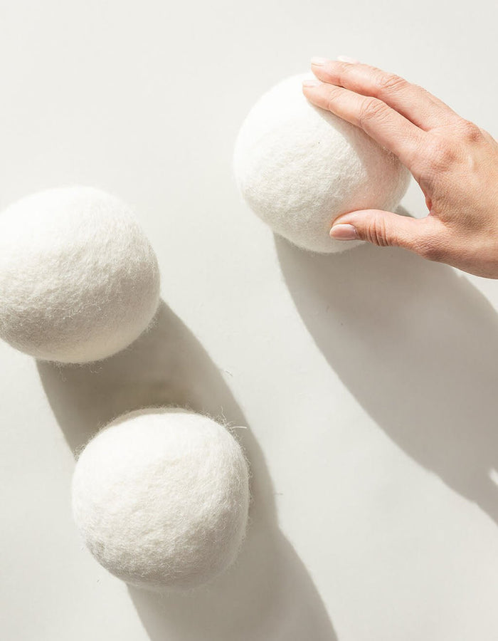 A hand reaches for one of three large Cozy Earth Wool Dryer Balls, displayed on a smooth, light background with soft shadows.|Size:Regular