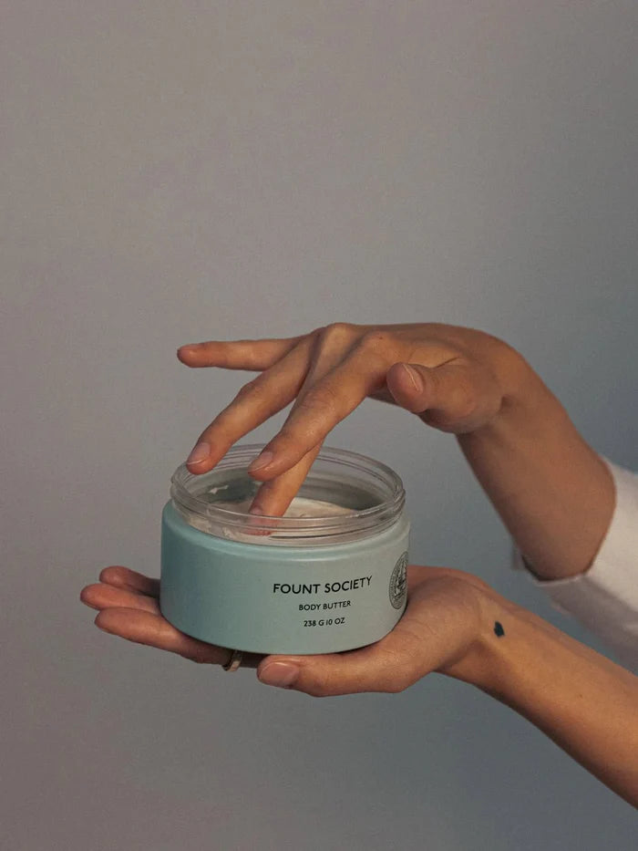 A woman holds Fount Society Body Butter in her hand and dips one finger into the lotion.